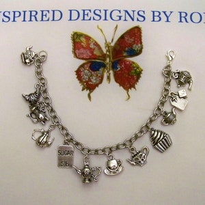 Könnte beinhalten: Ein silbernes Charm-Armband mit verschiedenen Charms zum Thema Tee, darunter Teekannen, eine Teetasse und ein Cupcake. Über dem Armband befindet sich eine dekorative Schmetterlingsbrosche. Der Text "SUGAR 5lb" befindet sich auf einem der Charms. Der Text "INSPIRED DESIGNS BY ROBIN" steht oben.