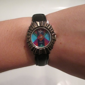 Puede incluir: Un reloj de pulsera de cuero negro con un diseño geométrico colorido en la esfera. La esfera del reloj está rodeada por un marco de metal plateado.