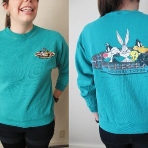 May include: Teal blue sweatshirt with a graphic of Bugs Bunny, Daffy Duck, Tweety Bird, and Sylvester the Cat on the back. The front of the sweatshirt has a small graphic of the Looney Tunes characters. The sweatshirt says "Looney Tunes" on the back.