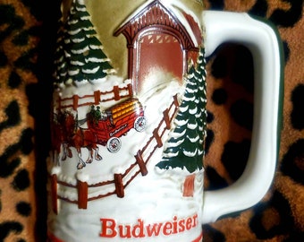 May include: White ceramic Budweiser beer stein featuring a winter scene. The stein depicts a red wagon pulled by two horses, a covered bridge, and snow-covered pine trees. The stein has green and red trim and the word "Budweiser" in red lettering.