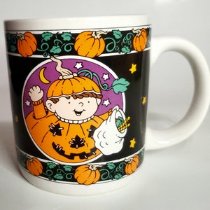 May include: A white ceramic mug with a black background and a colorful illustration of a child dressed as a pumpkin for Halloween. The child is holding a bag of candy and there are pumpkins and stars around the child.