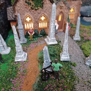 May include: A miniature village scene with a small church and a graveyard with several gray stone obelisks. There are small figures of people in the scene.