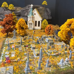以下が含まれることがあります： 小さな白い教会、十字架、そして多くの墓石がある墓地のミニチュアモデル。墓地は秋の落ち葉で覆われています