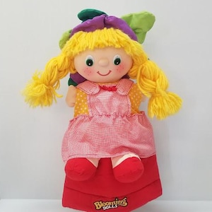 May include: A Blooming Dolls plush doll with yellow yarn hair in pigtails, a purple and green hat, and a red and white gingham dress. The doll has a smiling face and red shoes. The base is red with the words "Blooming Dolls".
