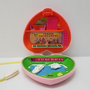 May include: A vintage Polly Pocket playset in a heart-shaped case, open to reveal a tennis tournament scene and a yacht race scene. The case is pink and orange with a yellow cord and white handle.