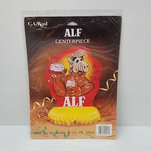 May include: ALF centerpiece in a plastic package. The centerpiece features a cartoon image of ALF holding a cake, with the text "Let's Party!" and "ALF". The package is 8 inches wide and 11 inches tall.