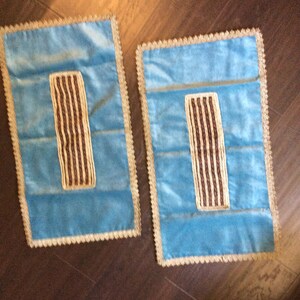 May include: Two rectangular, light blue velvet decorative pillows with a gold trim and a brown and gold striped design in the center. The pillows are placed on a dark brown wooden surface.