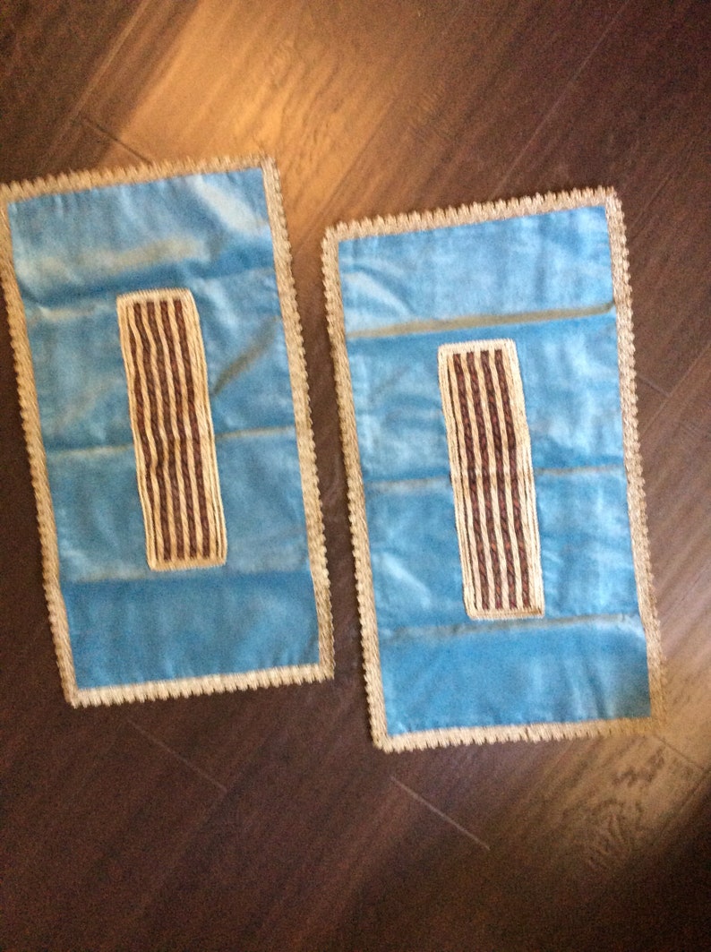 May include: Two rectangular, light blue velvet decorative pillows with a gold trim and a brown and gold striped design in the center. The pillows are placed on a dark brown wooden surface.