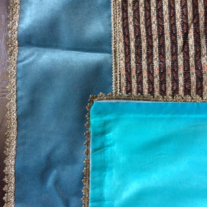May include: An overhead shot of three fabric swatches. One is a teal blue velvet, another is a striped fabric with brown, beige, and white vertical stripes, and the third is a bright turquoise satin. All three fabrics are trimmed with gold lace.
