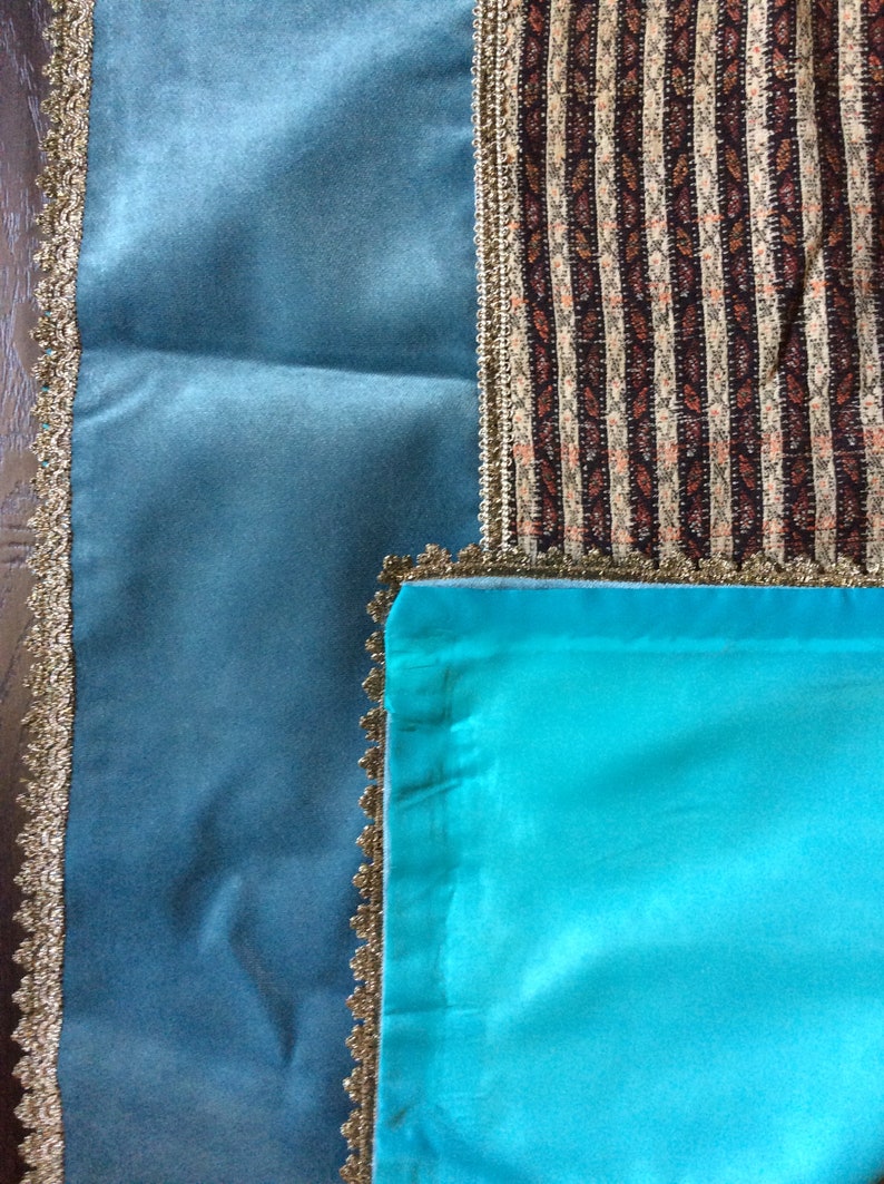 May include: An overhead shot of three fabric swatches. One is a teal blue velvet, another is a striped fabric with brown, beige, and white vertical stripes, and the third is a bright turquoise satin. All three fabrics are trimmed with gold lace.