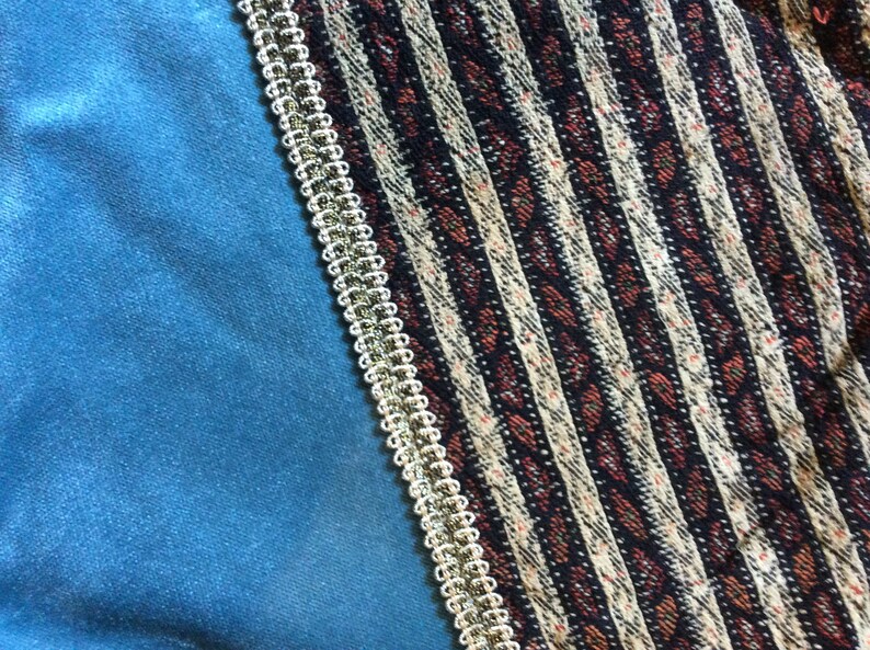 May include: A close-up of a textile with a teal velvet section on the left, a patterned section on the right, and a gold and white trim separating the two. The patterned section has vertical stripes in black, white, and red.