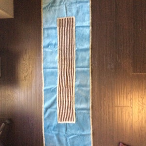 May include: A rectangular, light blue table runner with a central, vertical panel of brown and white stripes. The runner is trimmed with a gold-colored border and is displayed on a dark brown wooden surface.