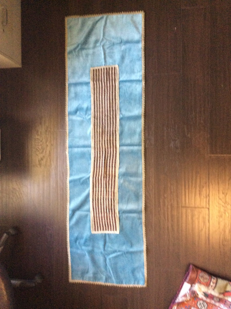 May include: A rectangular, light blue table runner with a central, vertical panel of brown and white stripes. The runner is trimmed with a gold-colored border and is displayed on a dark brown wooden surface.