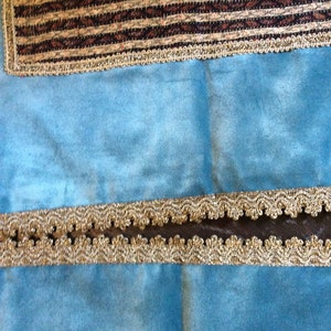 May include: Close-up of decorative trim on a light blue fabric. The trim features a gold-colored, scalloped edge with a dark brown center. A rectangular patch of fabric with a brown and tan striped pattern is also visible.
