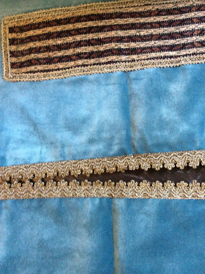May include: Close-up of decorative trim on a light blue fabric. The trim features a gold-colored, scalloped edge with a dark brown center. A rectangular patch of fabric with a brown and tan striped pattern is also visible.