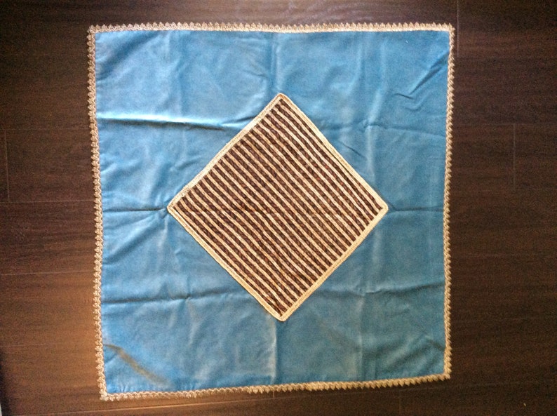 May include: A square, light blue pillow with a central diamond-shaped design. The diamond features brown and beige stripes, and the pillow has a decorative, beige trim around the edges. The pillow is likely for home decor.