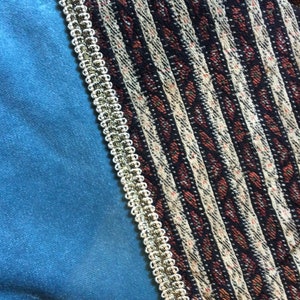 May include: A close-up of a textile with a teal blue velvet fabric on the left, a patterned fabric on the right, and a gold and white trim separating the two fabrics. The patterned fabric has vertical stripes in black, white, and red.