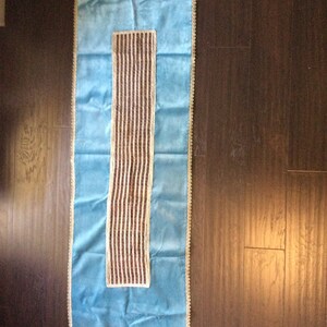 May include: A rectangular, light blue table runner with a central, vertical panel of brown and cream stripes. The edges are trimmed with a cream-colored, scalloped border. The runner is displayed on a dark wood surface.