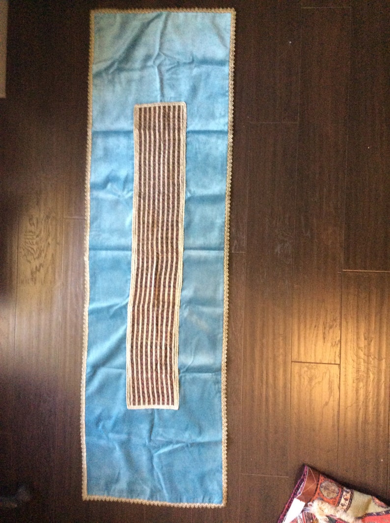 May include: A rectangular, light blue table runner with a central, vertical panel of brown and cream stripes. The edges are trimmed with a cream-colored, scalloped border. The runner is displayed on a dark wood surface.