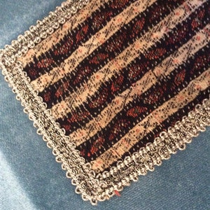 May include: Close-up of a decorative fabric piece with a striped pattern in black, cream, and red. The edges are trimmed with a silver, beaded trim. The fabric is set against a light blue background.