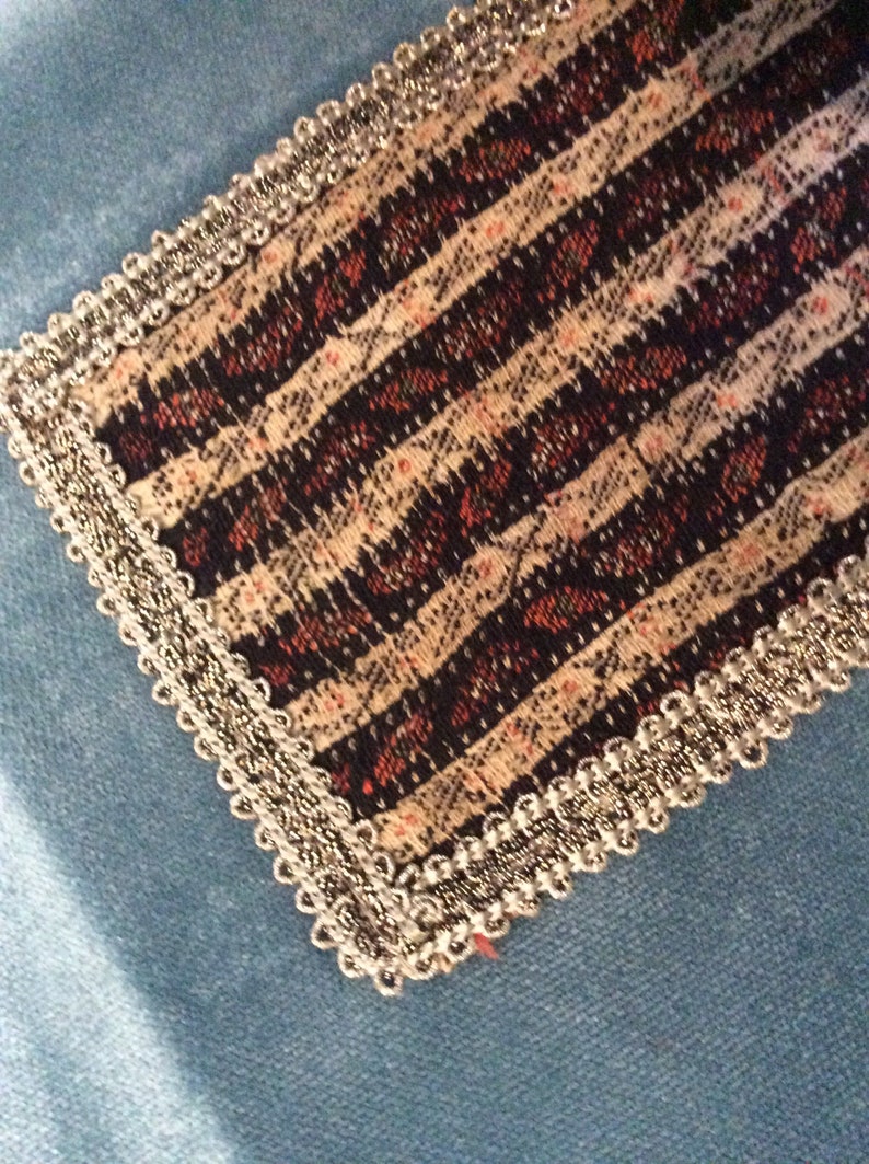 May include: Close-up of a decorative fabric piece with a striped pattern in black, cream, and red. The edges are trimmed with a silver, beaded trim. The fabric is set against a light blue background.