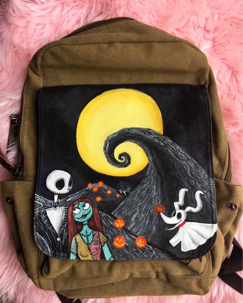 Puede incluir: Una mochila de lona marr&oacute;n con un parche de lona negra que presenta una escena pintada a mano de Pesadilla antes de Navidad. La escena incluye a Jack Skellington, Sally y Zero, con una luna amarilla y una colina en espiral en el fondo.