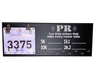 May include: Black chalkboard with a white inscription "PR Two little letters that make every runner smile".  The board has hooks for hanging race bibs and medals. A race bib with the number 3375 is hanging on the board.