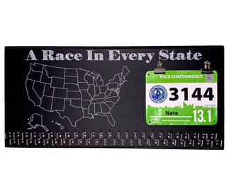 May include: A black chalkboard with a white outline of the United States map. The text "A Race In Every State" is written at the top. A green and white race bib with the number "3144" and the text "Race Chattanooga" is attached to the board with a silver clip.