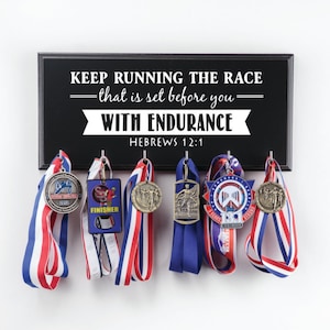 Puede incluir: Un soporte de madera negro para medallas con el texto "KEEP RUNNING THE RACE -that is set before you- WITH ENDURANCE HEBREWS 12:1" y varias medallas colgadas.