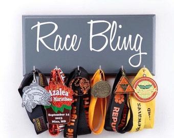 May include: A gray wooden medal display rack with the words "Race Bling" written in white. The rack has hooks for hanging medals and ribbons. There are seven medals and ribbons hanging on the rack, including a silver medal with a fish, a red medal with a flower, a black medal with a tiger, a gold medal with a number, a yellow medal with a triangle, a yellow medal with a green leaf, and a yellow medal with a red paw print.