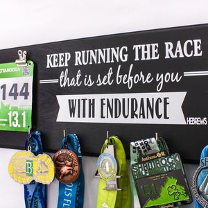 May include: Black wooden sign with the text "KEEP RUNNING THE RACE - that is set before you - WITH ENDURANCE = HEBREWS 12:1". The sign has hooks for hanging medals and a clip for a race bib. A green race bib with the number 3144 is clipped to the sign. Several medals are hanging from the hooks.