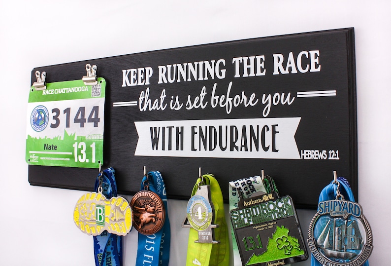 May include: Black wooden sign with the text "KEEP RUNNING THE RACE - that is set before you - WITH ENDURANCE = HEBREWS 12:1". The sign has hooks for hanging medals and a clip for a race bib. A green race bib with the number 3144 is clipped to the sign. Several medals are hanging from the hooks.