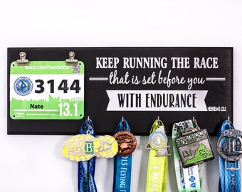 May include: Black wooden sign with the text "KEEP RUNNING THE RACE - that is set before you - WITH ENDURANCE HEBREWS 12:1". A green and white race bib with the number 3144 and the text "RACE CHATTANOOGA" is attached to the sign.  Five medals are hanging from hooks on the sign.