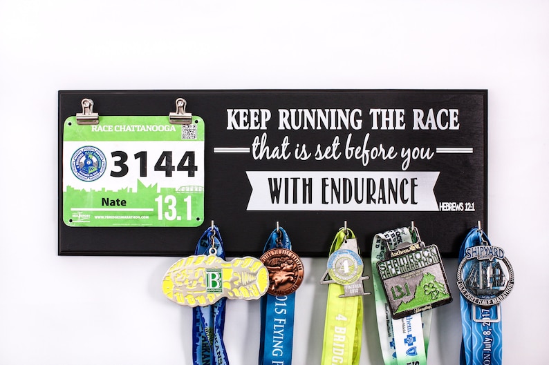 May include: Black wooden sign with the text "KEEP RUNNING THE RACE - that is set before you - WITH ENDURANCE HEBREWS 12:1". A green and white race bib with the number 3144 and the text "RACE CHATTANOOGA" is attached to the sign.  Five medals are hanging from hooks on the sign.