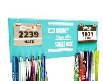 May include: A turquoise wooden medal display rack with two race bibs, one with the number 2239 and the other with the number 1971. Both bibs have the name "Nate" printed on them. The rack has a white inscription that reads "Every journey begins with a single step".  The rack has hooks for hanging medals, and there are several colorful medals hanging from the hooks.