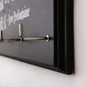 May include: Black wooden wall-mounted coat rack with silver hooks. The rack has the text "The best things in life are the small things -  Jose  Prefontaine" written on it.