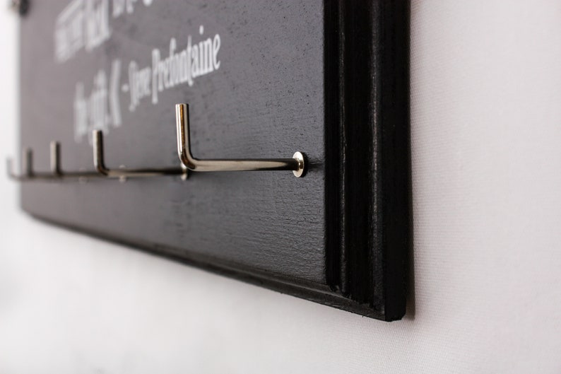 May include: Black wooden wall-mounted coat rack with silver hooks. The rack has the text "The best things in life are the small things -  Jose  Prefontaine" written on it.