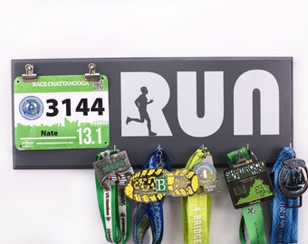 May include: A gray wooden medal display with the word "RUN" in white letters. The display has hooks for hanging medals and a clip for holding a race bib. The race bib is green and white and has the number "3144" on it. The text "RACE CHATTANOOGA" and "Nate - 13.1" are also on the bib.