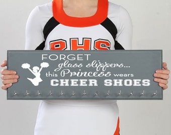 May include: Gray wooden sign with the text "FORGET glass slippers... this Princess wears CHEER SHOES" and a white silhouette of a cheerleader with pom-poms. The sign has eight hooks for hanging items.