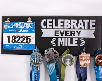 May include: A black wooden medal display with the text "Celebrate Every Mile" in silver lettering. The display has hooks for hanging medals and a clip for holding a race bib. A blue race bib with the number 18225 is clipped to the display. Several medals are hanging from the hooks, including a silver medal with the word "Ironman" on it.