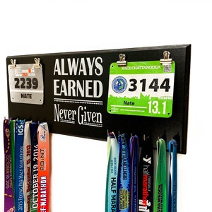 May include: Black wooden sign with the text "Always Earned Never Given" and two race bibs attached with clips. The bibs are green and white with the numbers "2239" and "3144".  The sign has hooks for hanging medals.  There are several colorful medals hanging on the hooks.
