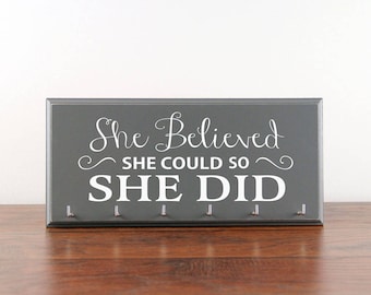 May include: Gray wooden sign with the text "She Believed She Could So She Did" in white lettering. The sign has 7 hooks for hanging medals or other items.