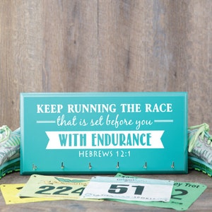 Puede incluir: Un letrero de madera azul turquesa con el texto "KEEP RUNNING THE RACE -that is set before you- WITH ENDURANCE HEBREWS 12:1" en letras blancas. El letrero tiene cinco ganchos para colgar llaves u otros art&iacute;culos. En primer plano hay dos pares de zapatillas de correr verdes y tres dorsales de carrera con n&uacute;meros.