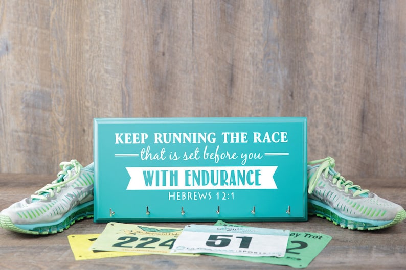 Puede incluir: Un letrero de madera azul turquesa con el texto "KEEP RUNNING THE RACE -that is set before you- WITH ENDURANCE HEBREWS 12:1" en letras blancas. El letrero tiene cinco ganchos para colgar llaves u otros art&iacute;culos. En primer plano hay dos pares de zapatillas de correr verdes y tres dorsales de carrera con n&uacute;meros.