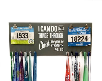 May include: A gray wooden medal and race bib holder with the text "I can do all things through Christ who gives me strength Phil. 4:13". The holder has hooks for hanging medals and clips for displaying race bibs. There are several medals and bibs displayed on the holder.