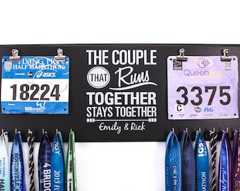 May include: Black wooden sign with the text "The couple that runs together stays together" with the names "Emily & Rick" below. Two race bibs are attached to the sign with clips, one blue with the number "18224" and one purple with the number "3375".  Below the sign are several colorful running medals hanging on hooks.