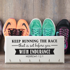 Puede incluir: Un zapatero de madera blanco con ganchos y el texto "KEEP RUNNING THE RACE - that is set before you - WITH ENDURANCE HEBREWS 12:1". Hay seis pares de zapatillas para correr en el fondo.