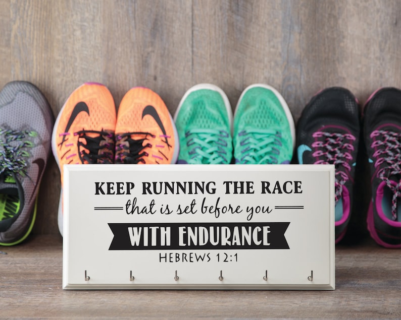 Puede incluir: Un zapatero de madera blanco con ganchos y el texto "KEEP RUNNING THE RACE - that is set before you - WITH ENDURANCE HEBREWS 12:1". Hay seis pares de zapatillas para correr en el fondo.