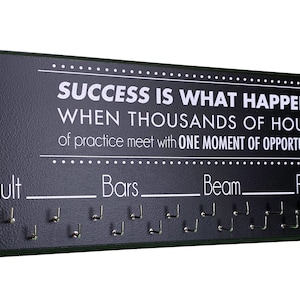 May include: Black wooden wall-mounted medal display with hooks for hanging medals. The display has the text "SUCCESS IS WHAT HAPPENS WHEN THOUSANDS OF HOURS of practice meet with ONE MOMENT OF OPPORTUNITY" printed on it. The hooks are labeled "Vault", "Bars", "Beam", and "Floor".