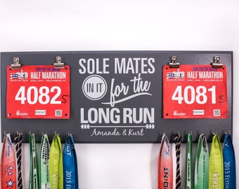 May include: A gray wooden medal display with the text "Sole Mates In It For The Long Run Amanda & Kurt". Two red race bibs are attached to the display with clips. The bibs have the numbers "4082" and "4081".  A row of colorful medals hangs below the bibs.